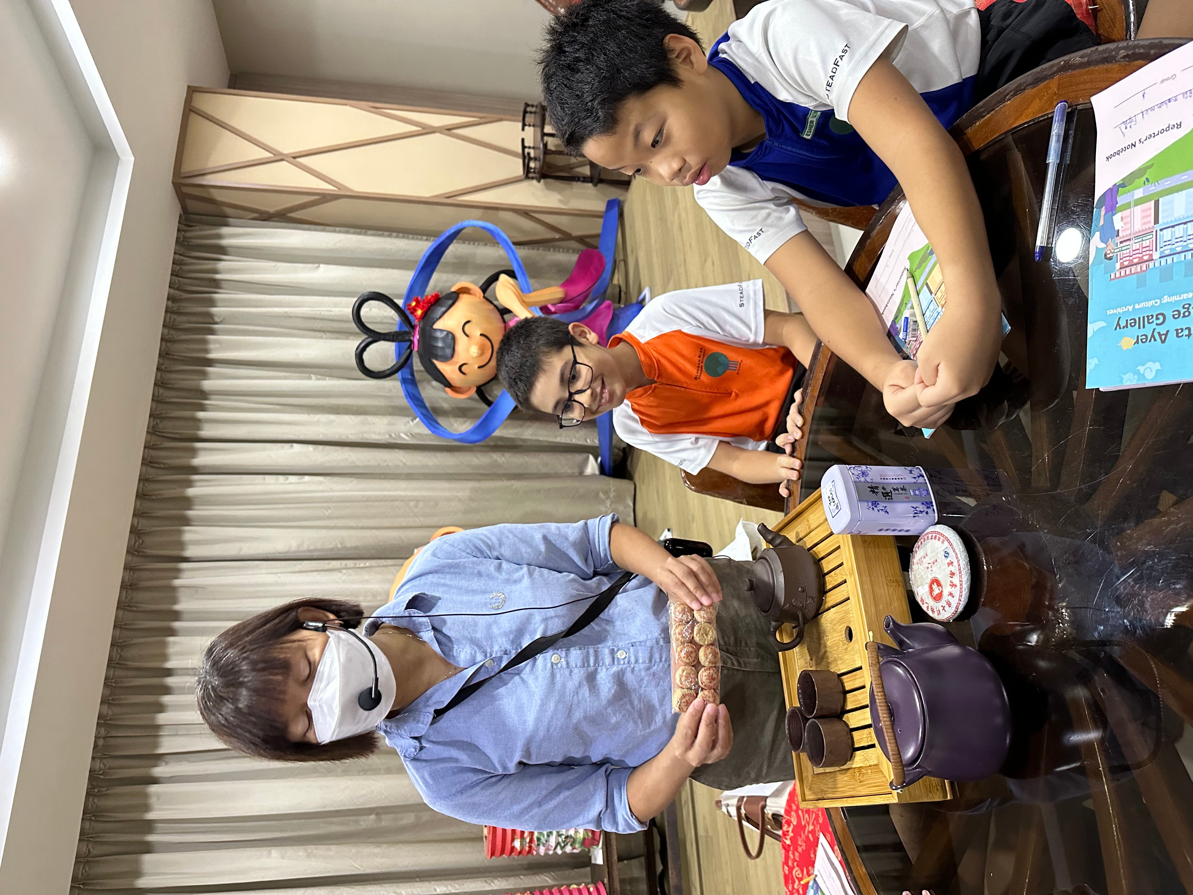 A masked woman with a headset shows tea items to two boys seated at a table with teapots and cups.